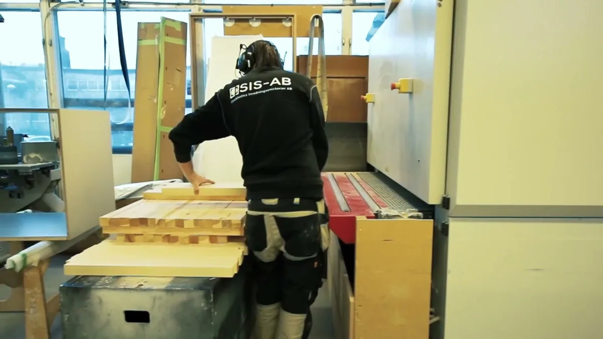 Snickare i SIS-AB-jacka och hörselkåpor vid en stapel trälister framför en maskin i verkstaden i Bandhagen
